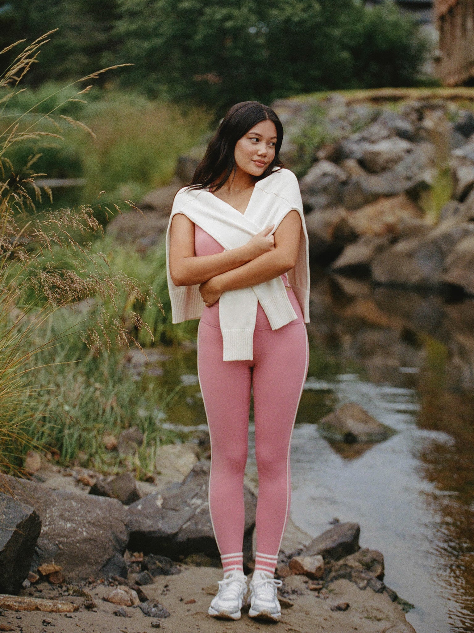 Woman in pink leggings and white top standing by a stream with rocks and greenery.