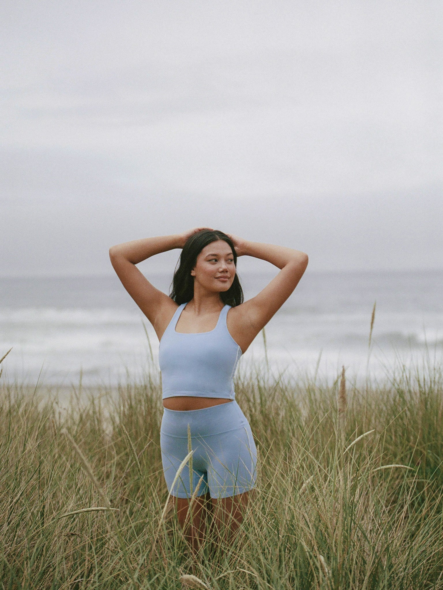 Woman in light blue athletic wear standing in tall grass with a cloudy sky background