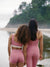 Two women in pink athletic wear standing back-to-back by a body of water with greenery.