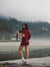Woman in maroon outfit standing on a beach with trees and fog in the background