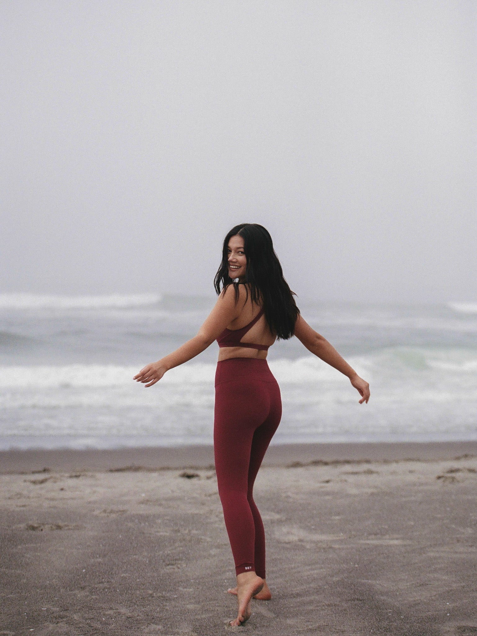 Woman in burgundy leggings and top standing on a beach with ocean waves in the background