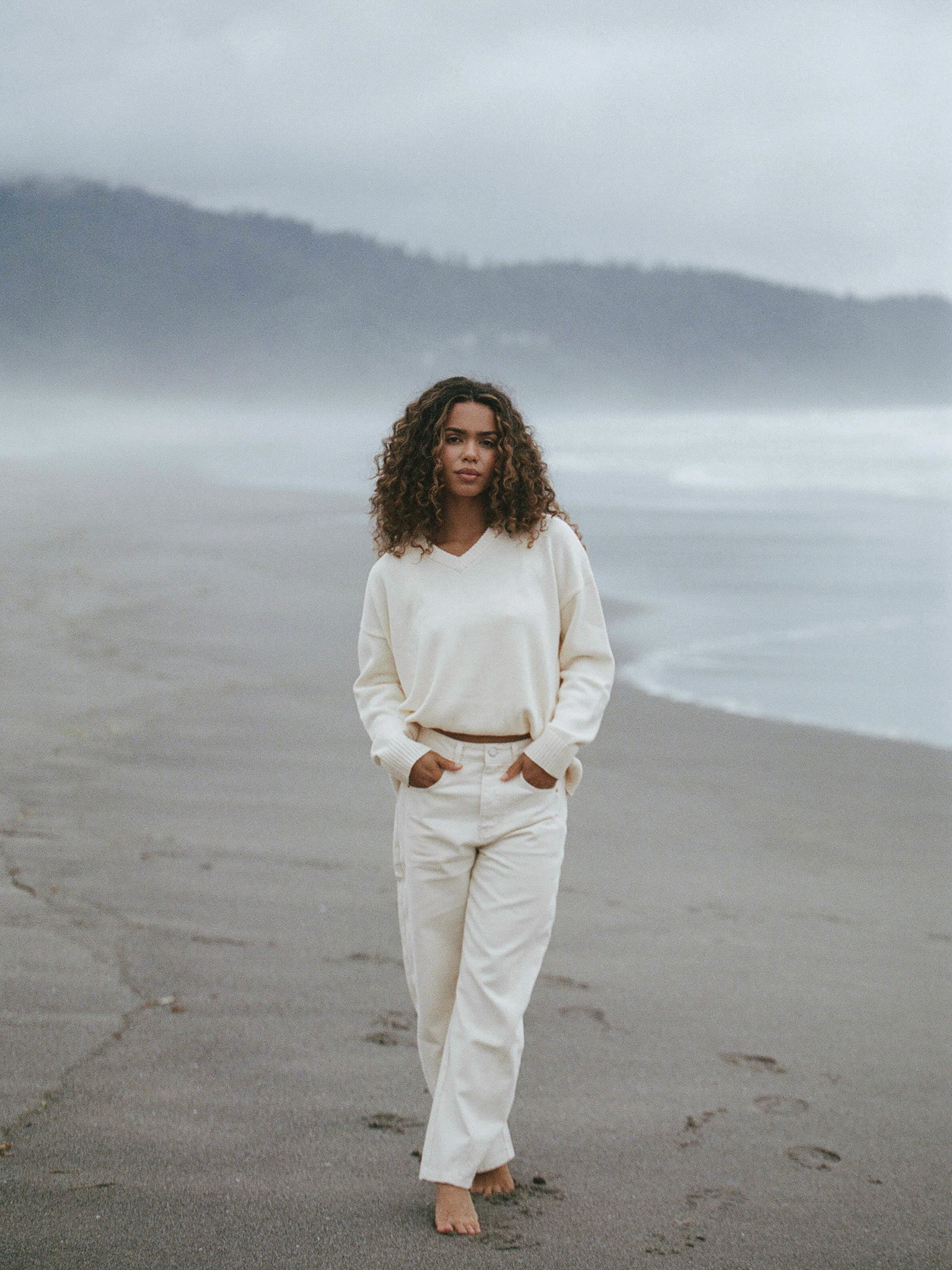 Person wearing a white outfit standing on a beach with foggy mountains in the background