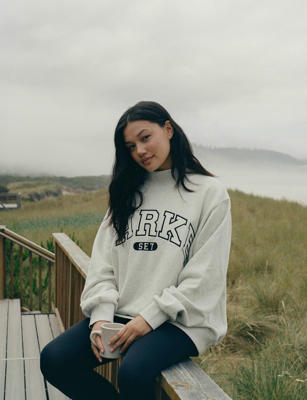 Woman sitting outdoors holding a mug, wearing a sweatshirt with text