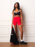 A woman stands against a white paneled wall wearing a black sports bra with white trim and bright red high-waisted shorts, holding a black jacket with red accents.