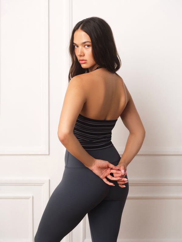A woman posing from behind wearing a black and gray striped tube top and dark gray leggings against a white paneled wall.