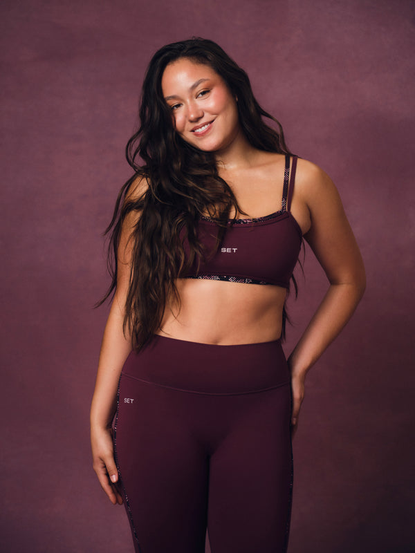 A woman modeling a matching burgundy activewear set consisting of a sports bra and high-waisted leggings, posed against a solid mauve studio backdrop. 