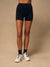 A model from the waist down wearing black high-waisted biker shorts, black crew socks, and white trail running shoes against a solid beige studio background.