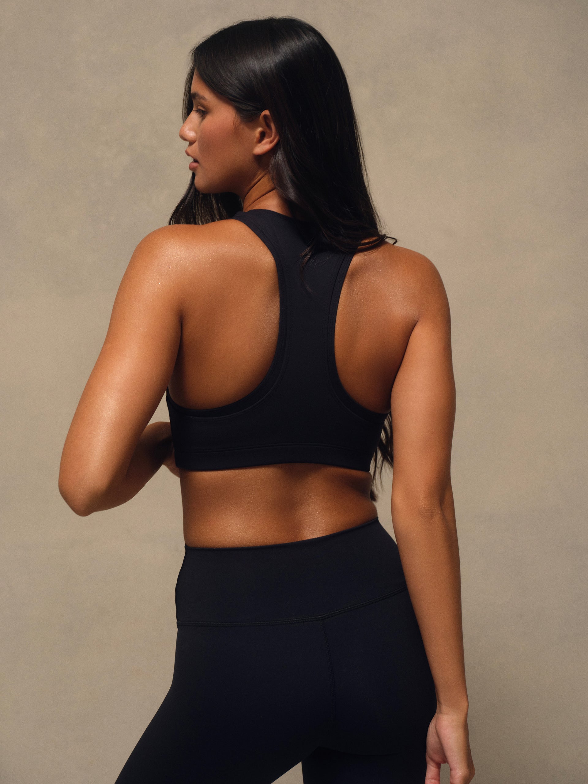 A woman shown from the back wearing a black sports bra and matching black leggings against a neutral beige studio background.