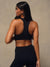 A woman shown from the back wearing a black sports bra and matching black leggings against a neutral beige studio background.