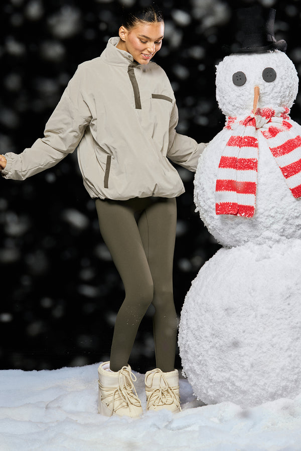 Person in winter clothing standing next to a snowman with a red scarf against a snowy background