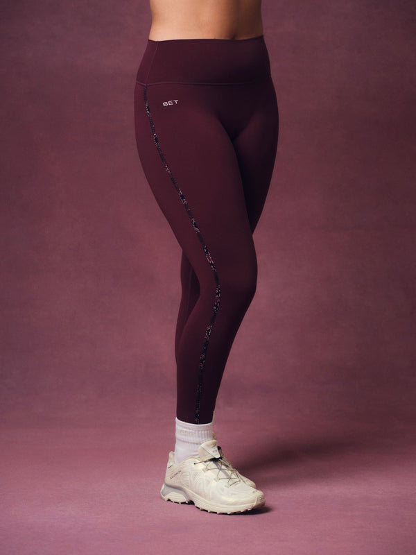 A person wearing high-waisted burgundy athletic leggings with a decorative side stripe and a small white 'SET' logo, paired with white socks and off-white sneakers, against a solid mauve studio backdrop.