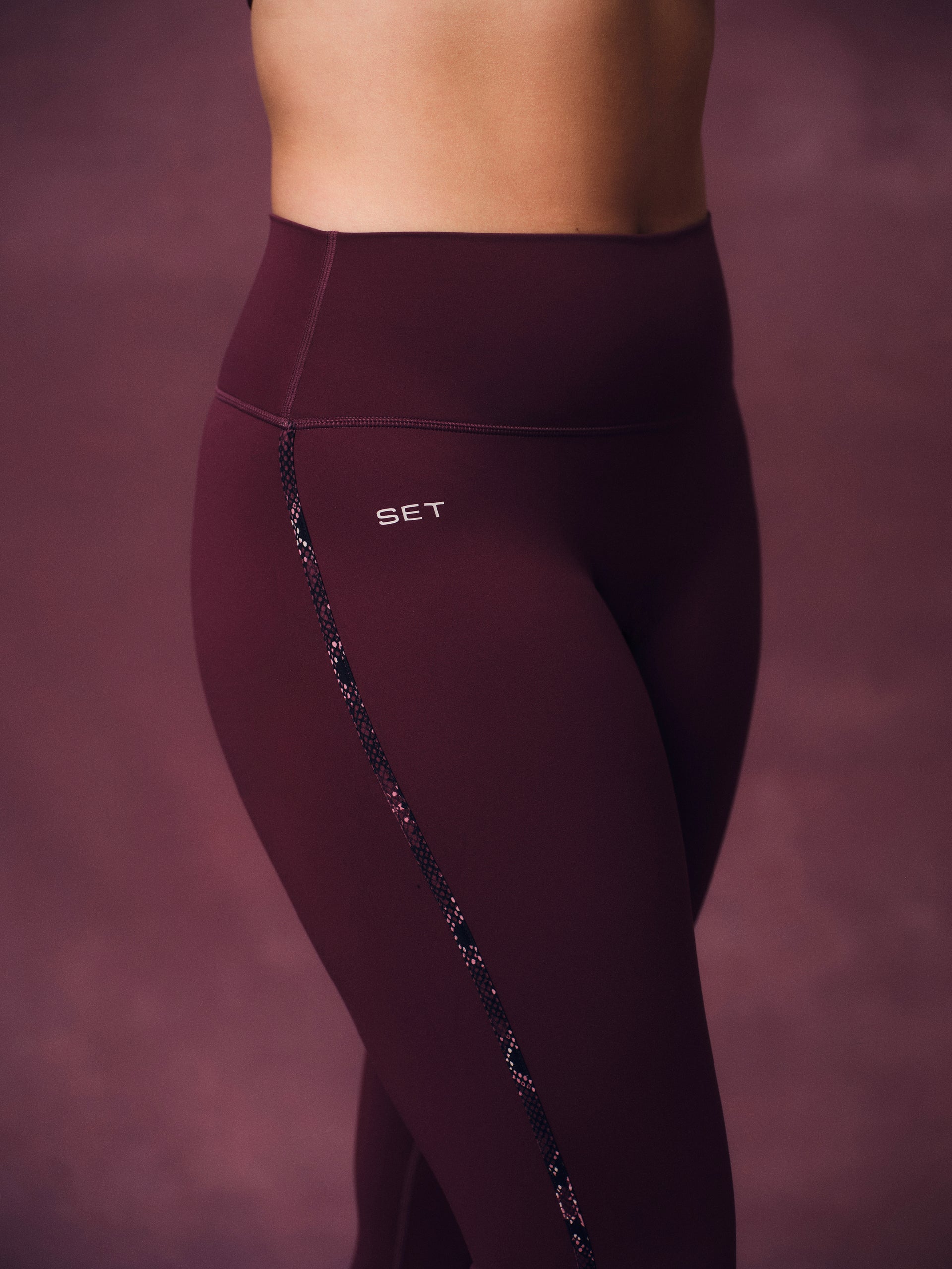 Close-up of a model wearing high-waisted burgundy leggings with a decorative snake skin detail running down the side and a small white SET logo on the hip, against a matching textured studio backdrop.