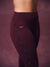 Close-up of a model wearing high-waisted burgundy leggings with a decorative snake skin detail running down the side and a small white SET logo on the hip, against a matching textured studio backdrop.