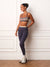 A woman modeling a matching activewear set consisting of a navy blue and beige striped sports bra and high-waisted navy blue leggings, standing against a white paneled wall.