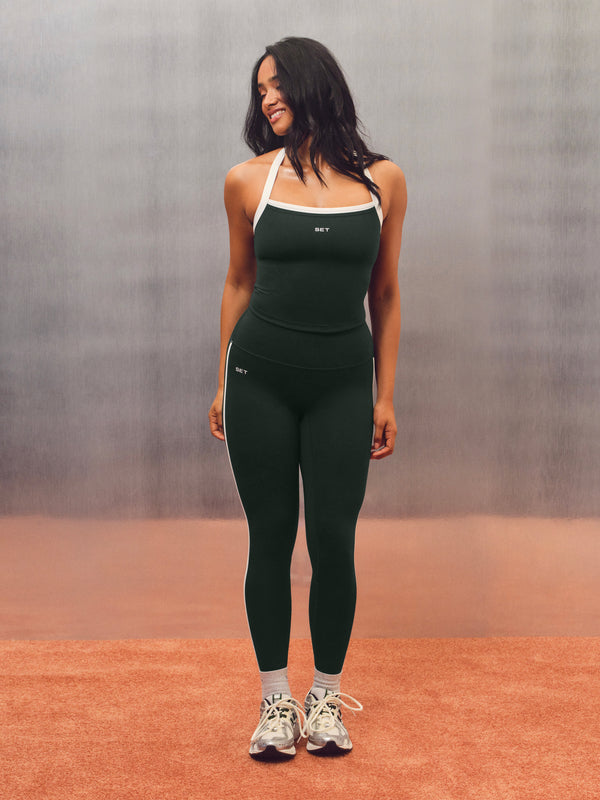 A woman modeling a matching black athletic set consisting of a tank top and leggings, featuring white trim and side stripes, standing on a terracotta-colored floor against a textured gray backdrop.  