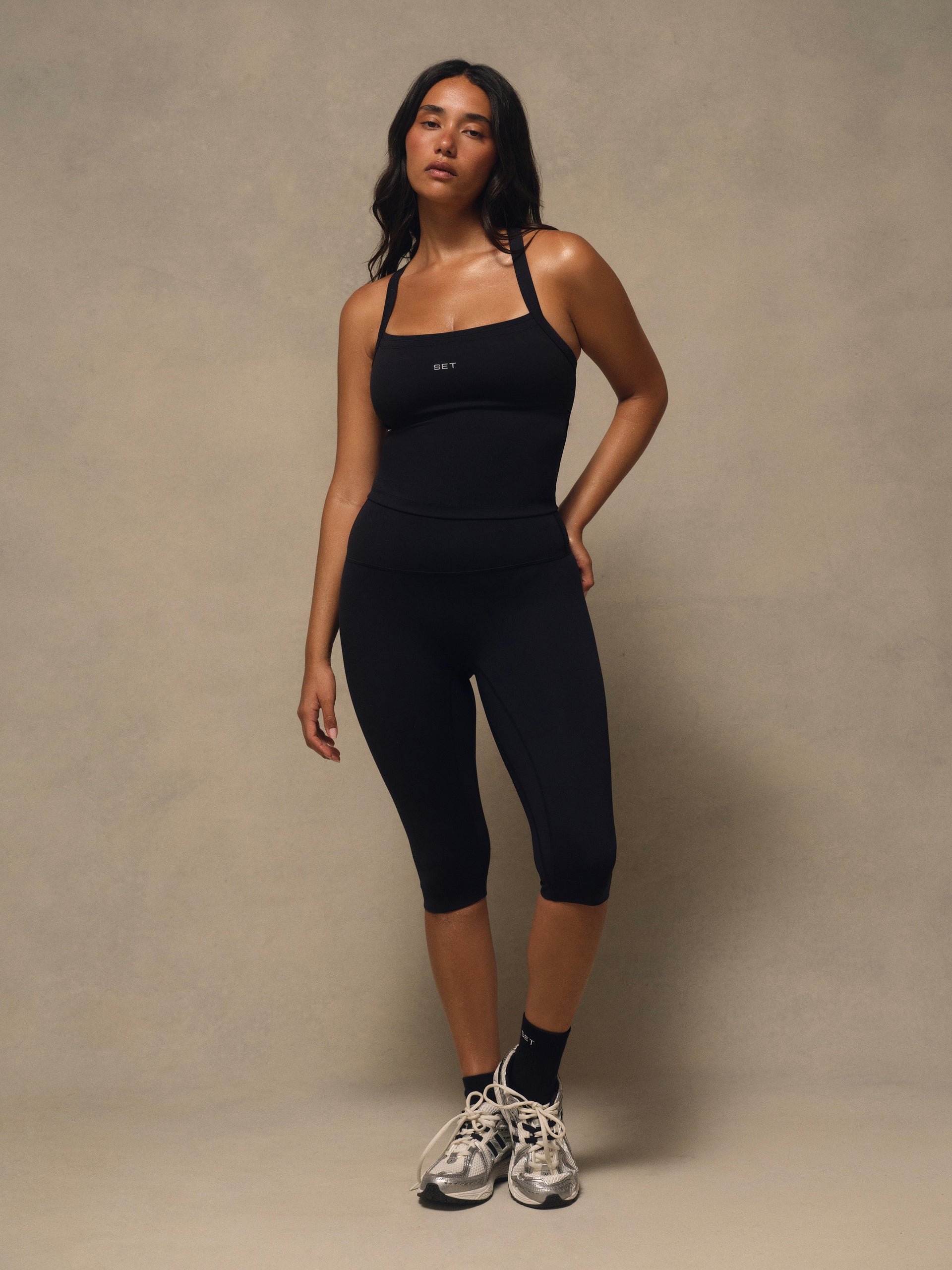 A woman modeling a black athletic tank top and matching black capri leggings against a neutral beige studio backdrop.