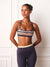 A woman modeling a navy blue sports bra with beige and white horizontal stripes, paired with dark navy leggings, standing against a white paneled wall.