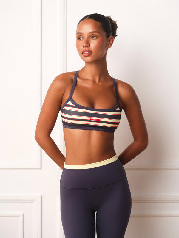 A woman modeling a navy blue sports bra with beige and white horizontal stripes, paired with dark navy leggings, standing against a white paneled wall.