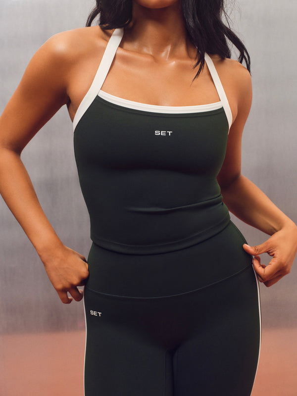 A woman wearing a dark forest green athletic tank top and matching leggings with white trim and a small white 'SET' logo, shown from the neck down against a gray studio backdrop. 