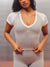 A woman wearing a light gray and white short-sleeve bodysuit with a V-neck, shown from the head down to the upper thighs. She is pulling the waistband slightly outward with both hands. <model-info>Alana wears a size M and is 5’8”, 28” waist, 35" bust.</model-info>
