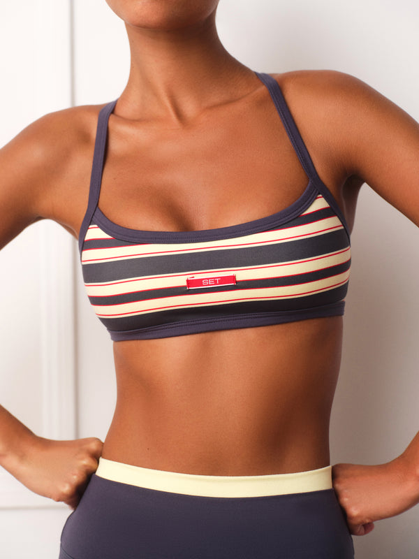 A close-up of a woman wearing a navy blue sports bra with cream and red horizontal stripes, featuring a small red rectangular logo patch on the front.