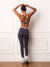 A woman is shown from behind wearing a matching athletic set consisting of a grey blue sports bra with beige stripes and high-waisted grey blue leggings with a beige waistband, standing against a white paneled wall.