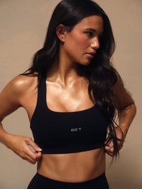 A woman wearing a black scoop-neck athletic sports bra with a small white 'SET' logo on the front, posed against a warm beige studio background. 