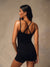 A woman shown from the back wearing a black strappy bodysuit with matching shorts, photographed against a textured warm gray studio backdrop.
