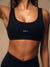 A close-up of a woman wearing a black scoop-neck athletic sports bra with the word 'SET' printed in white on the chest, paired with black bottoms against a warm beige background. <model-info>Lena wears a size XS and is 5’7”, 24½” waist, 31" bust.</model-info>