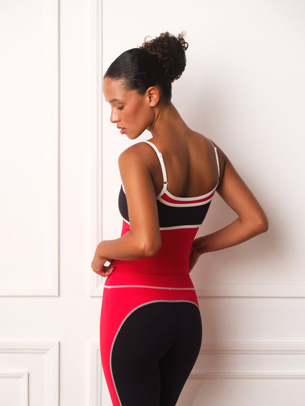 A woman wearing a red, black, and white athletic leotard stands in profile against a white paneled wall, showing the back of the garment.