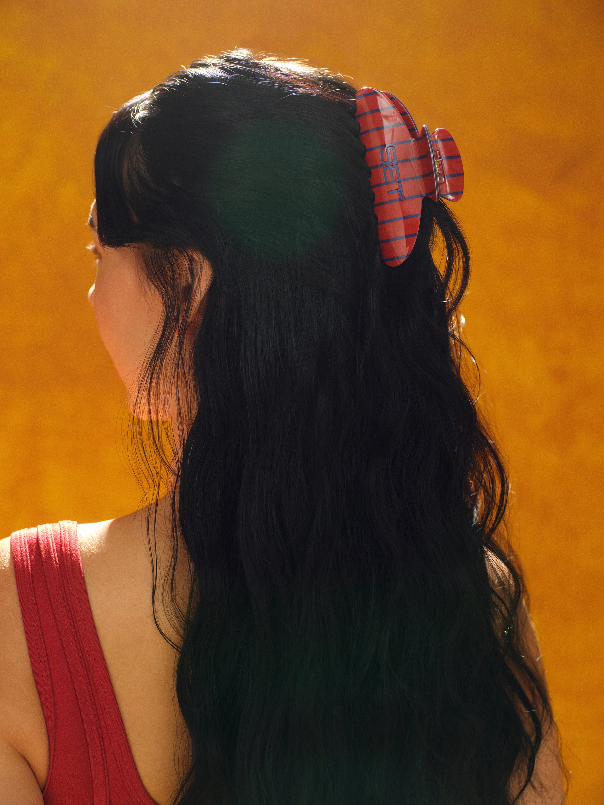 Person with long dark hair wearing a red and blue plaid hair clip against an orange background