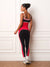 A woman stands with her back to the camera wearing a matching red, black, and white color-blocked activewear set consisting of a tank top and leggings, paired with white socks and gray running shoes, set against a white paneled wall.