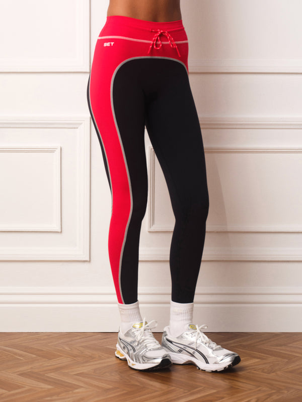 A person wearing high-waisted athletic leggings in red, black, and gray side panels, paired with white socks and white sneakers, standing against a white paneled wall.