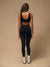 A woman is shown from behind wearing a black sports bra and matching black high-waisted leggings, standing against a solid beige studio backdrop.