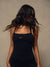 A woman with long, dark, wind-blown hair is shown from the back wearing a black athletic tank top with a small white 'SET' logo on the upper back, set against a neutral textured beige background.