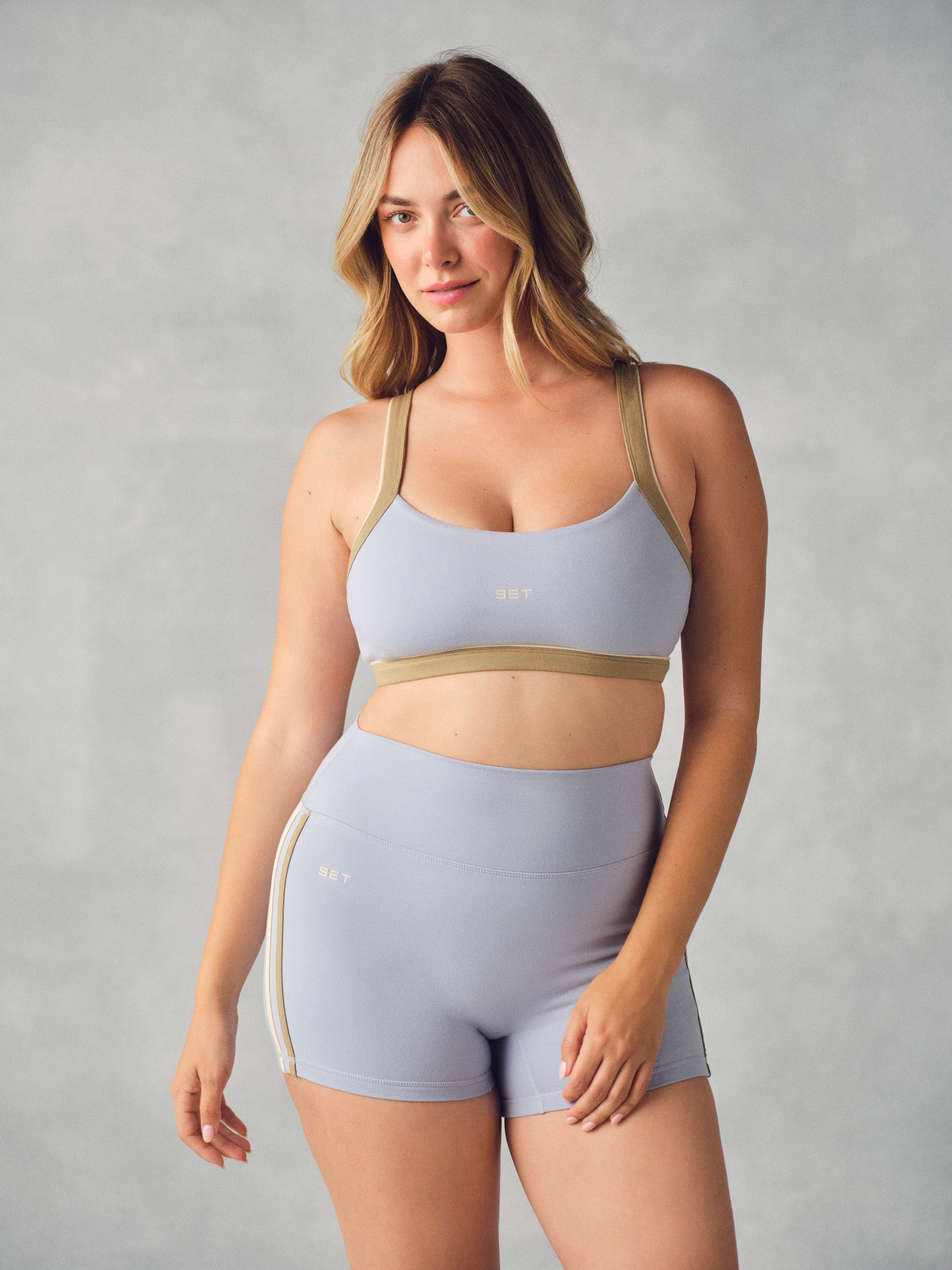 A woman modeling a matching light blue-gray activewear set consisting of a sports bra and high-waisted shorts, featuring beige contrast straps and side stripes, against a textured gray studio background. 
