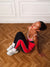 A woman sits on a wooden herringbone floor wearing a red, black, and white athletic one-piece jumpsuit, white socks, and white sneakers.