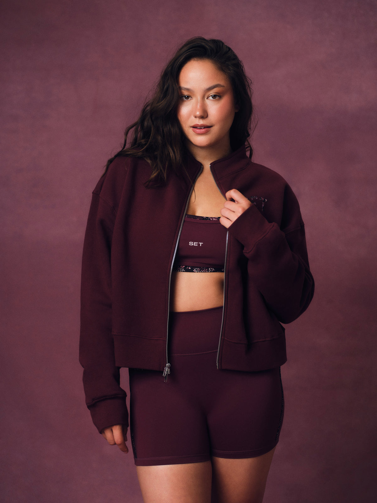 A woman modeling a matching burgundy activewear set consisting of a zip-up jacket, sports bra, and high-waisted shorts against a textured mauve-brown studio backdrop. <model-info> Christina wears a size L and is 5’9, 29½” waist, 36" bust.</model-info> 