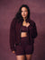 A woman modeling a matching burgundy activewear set consisting of a zip-up jacket, sports bra, and high-waisted shorts against a textured mauve-brown studio backdrop. <model-info> Christina wears a size L and is 5’9, 29½” waist, 36" bust.</model-info> 