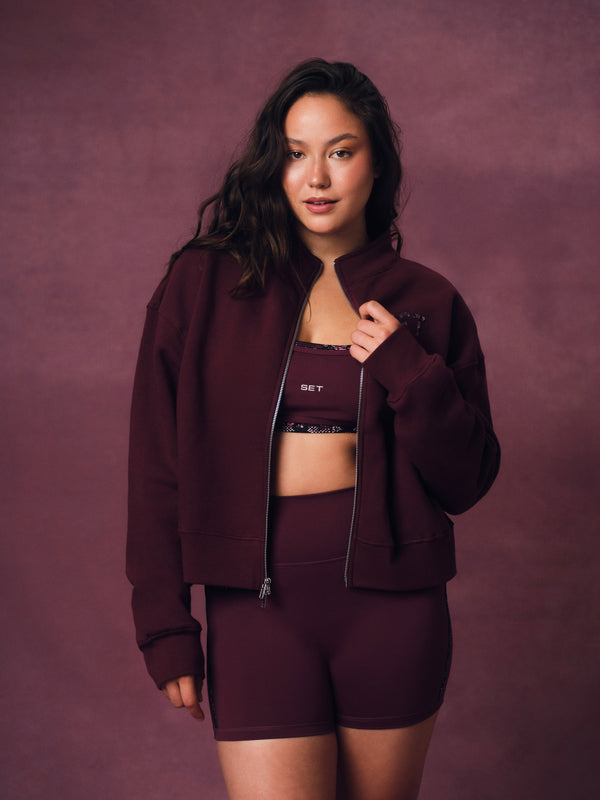 A woman modeling a matching burgundy activewear set consisting of a zip-up jacket, sports bra, and high-waisted shorts against a textured mauve-brown studio backdrop. 