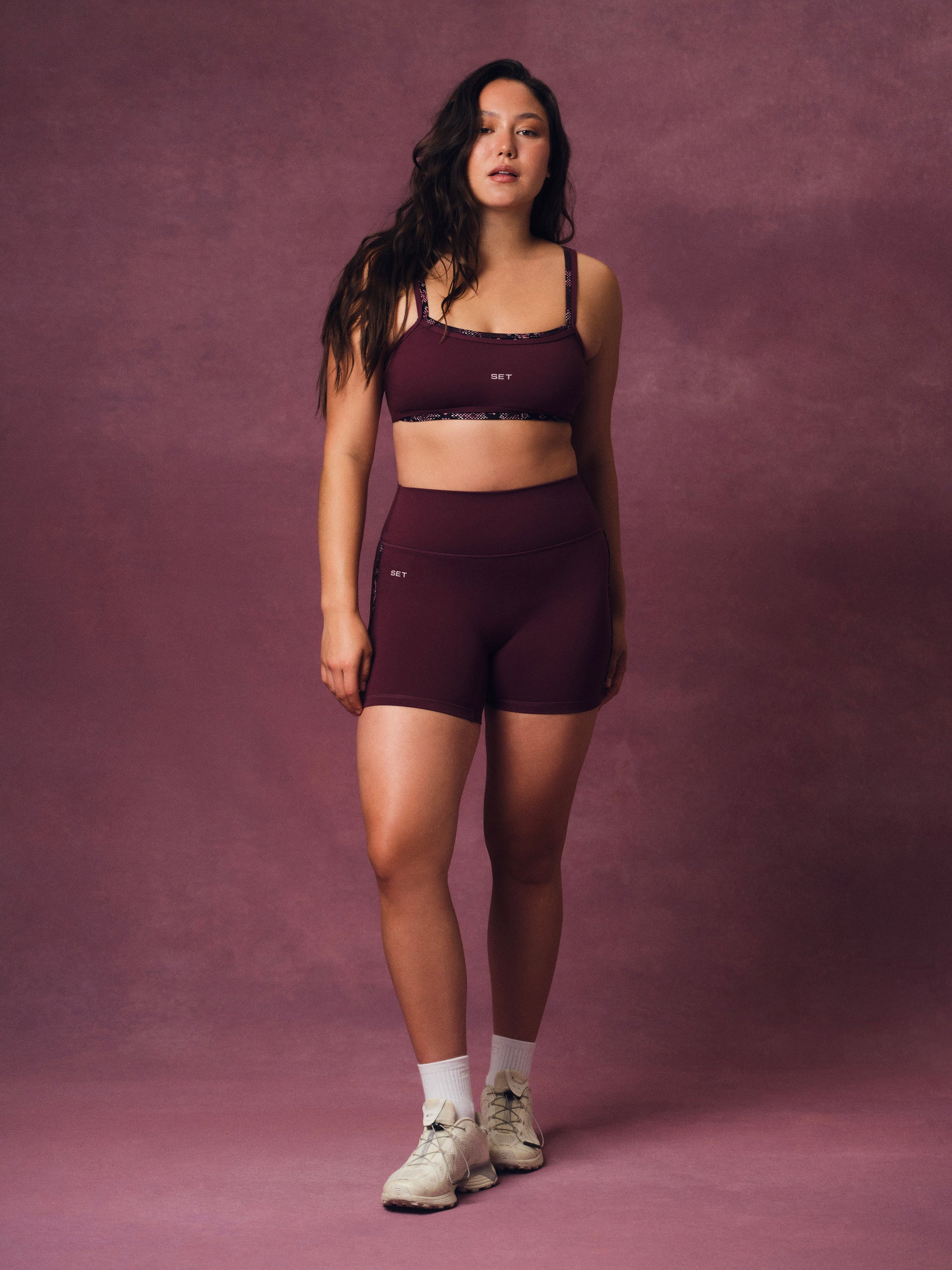 A woman modeling a matching burgundy activewear set consisting of a sports bra and high-waisted shorts, standing against a textured mauve studio backdrop.