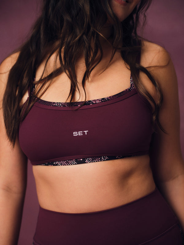 A close-up of a woman wearing a burgundy sports bra with black patterned trim and the brand name 'SET' printed in white on the front, against a matching burgundy background.