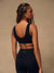 A woman shown from the back wearing a black sports bra and matching black leggings against a plain beige studio background. The leggings have a small white 'SET' logo on the waistband.