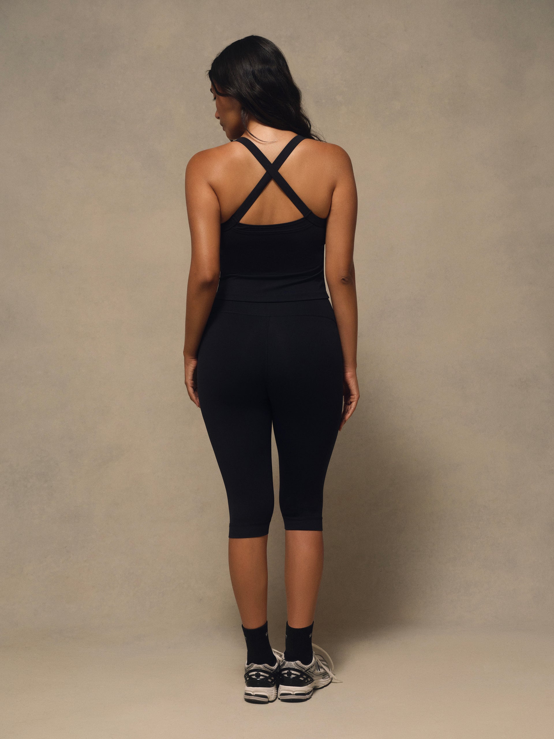 A woman is shown from behind wearing a black athletic tank top with crisscross straps and matching black capri leggings, standing against a neutral textured studio backdrop.