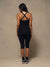 A woman is shown from behind wearing a black athletic tank top with crisscross straps and matching black capri leggings, standing against a neutral textured studio backdrop.