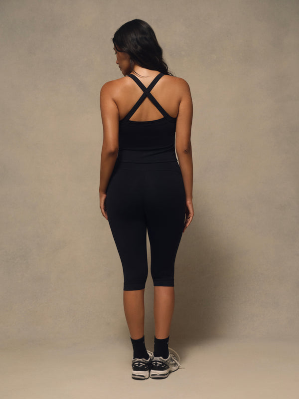 A woman is shown from behind wearing a black athletic tank top with crisscross straps and matching black capri leggings, standing against a neutral textured studio backdrop.