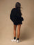 A woman is shown from behind wearing an oversized black crewneck sweatshirt, black shorts, black socks, and chunky white sneakers against a solid beige studio backdrop.