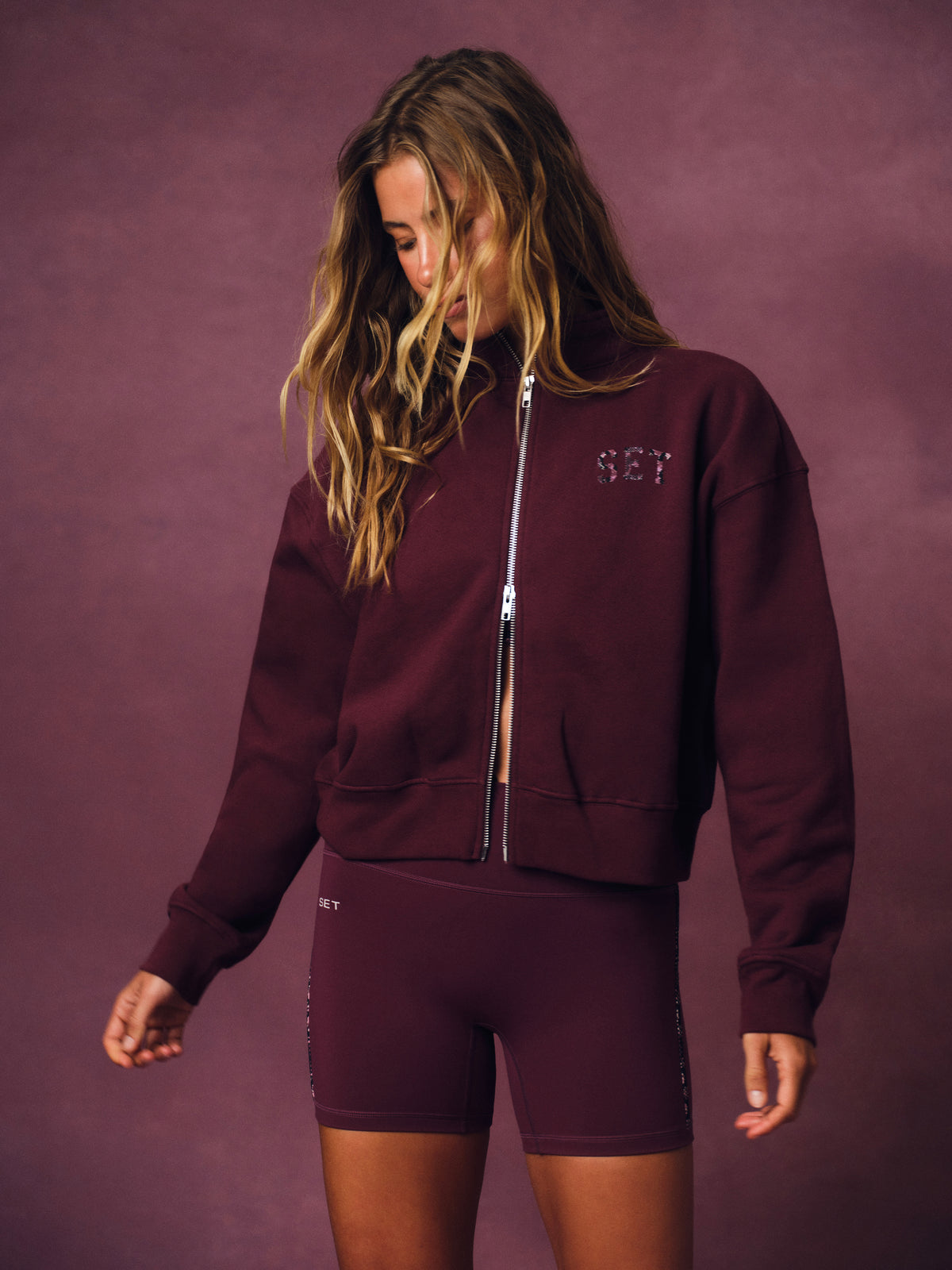 A woman modeling a matching burgundy zip-up sweatshirt and biker shorts set against a textured mauve studio backdrop. The sweatshirt features a silver zipper and a small silver 'SET' logo  on the left chest. 
