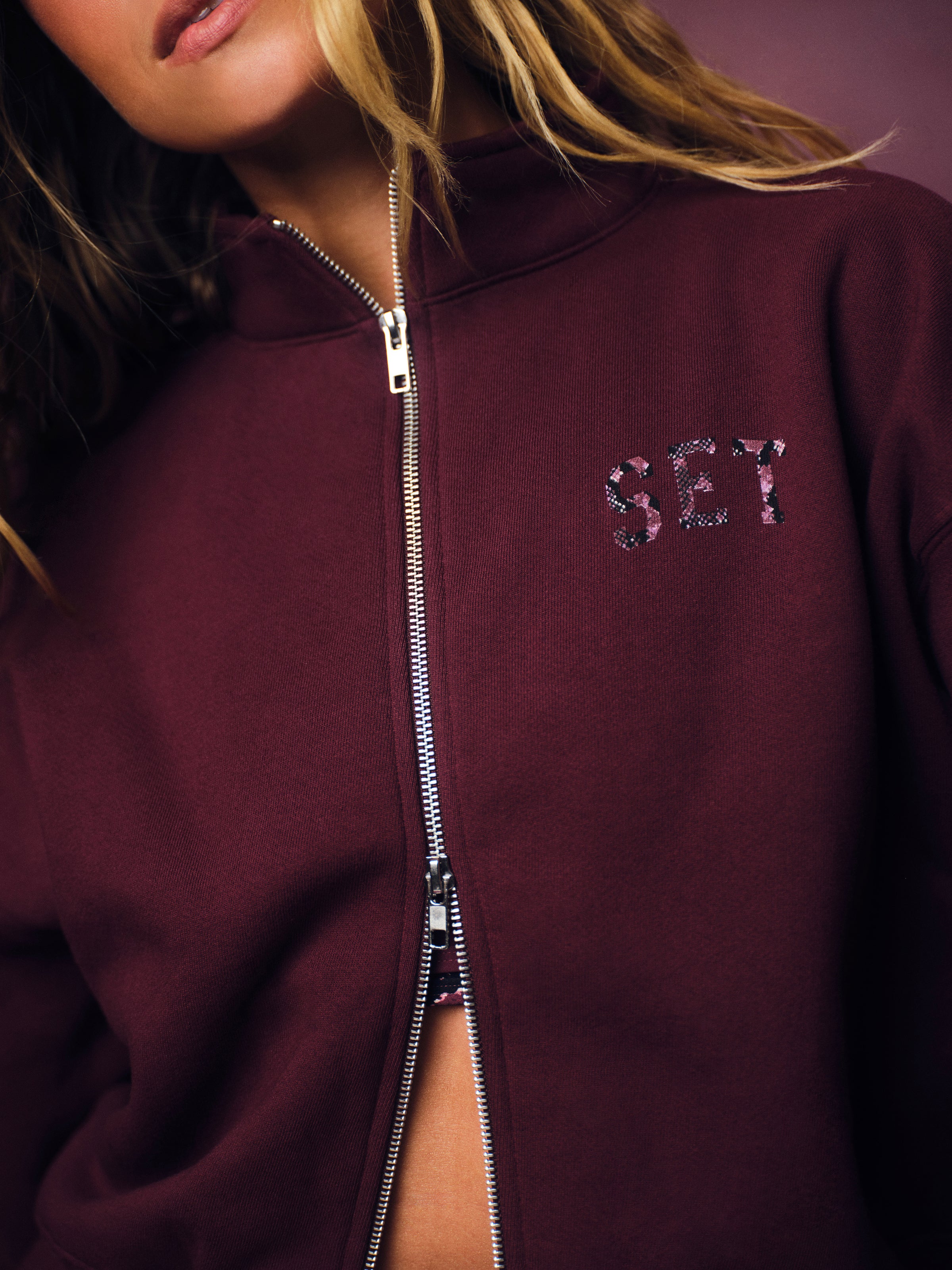 A close-up of a model wearing a burgundy zip-up hoodie with a silver zipper and 'SET' embroidered on the chest in a patterned thread.