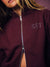 A close-up of a model wearing a burgundy zip-up hoodie with a silver zipper and 'SET' embroidered on the chest in a patterned thread.